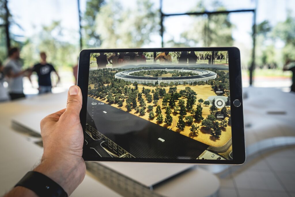 A person's hand holding a tablet that displays an augmented reality (AR) view of a circular building surrounded by trees, showcasing the innovative AR capabilities Fortum Digital Services provides for the real estate industry. The tablet overlays digital enhancements onto the real-world scene, exemplifying how AR can be used to offer immersive 3D tours and interactive visual content for real estate marketing.