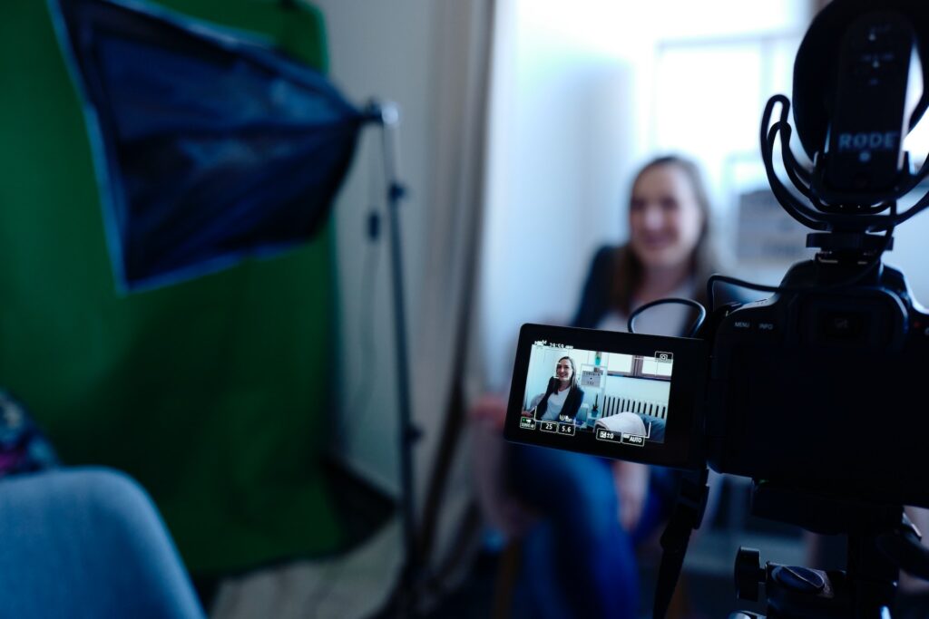 A behind-the-scenes view of a video marketing session by Fortum Digital Services, showcasing a digital camera focused on a smiling woman being interviewed in a professional setting. The camera's display screen highlights the interviewee, emphasizing Fortum Digital's commitment to leveraging personal stories and expert insights to enhance their real estate digital solutions.