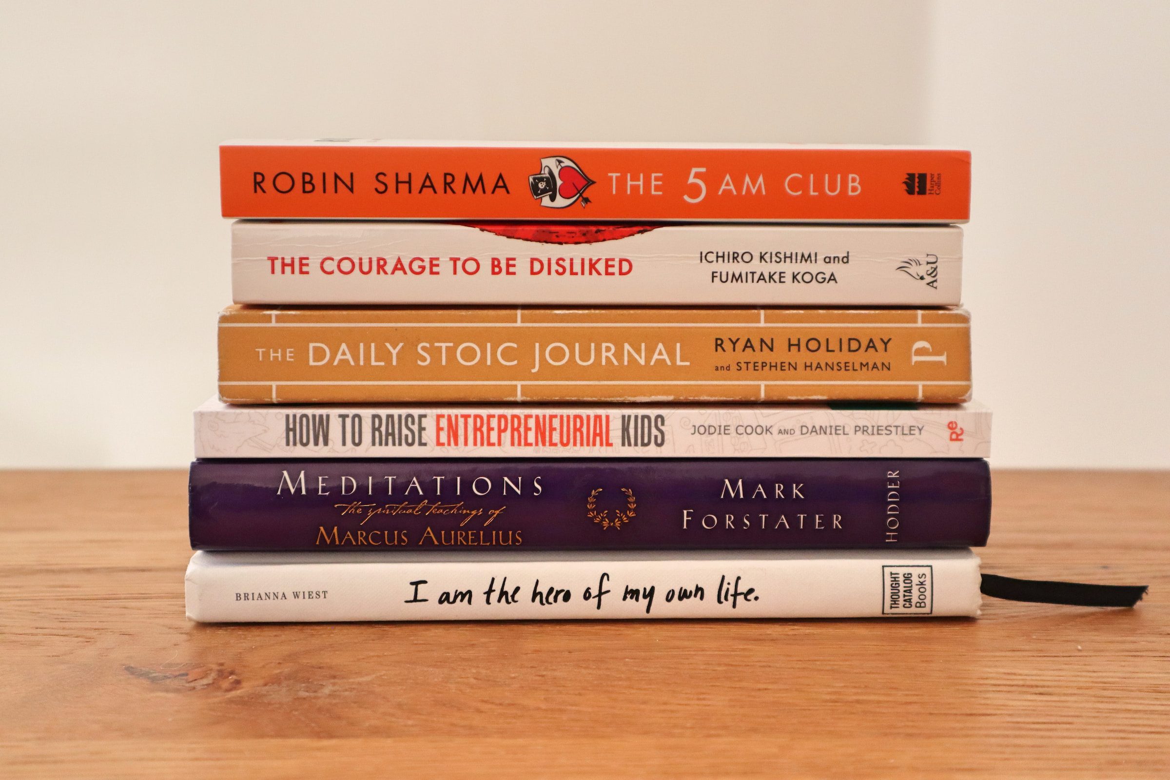 Assorted Stack of Business Books: Robin Sharma's The 5AM Club, Ichiro Kishimi and Fumitake Koga's The Courage To Be Disliked, Ryan Holiday's Daily Stoic Journal, Jodie Cook and Daniel Priestley's How To Raise Entrepreneurial Kids, Marcus Aurelius' Meditations, and Brianna Wiest's I am the hero of my life.